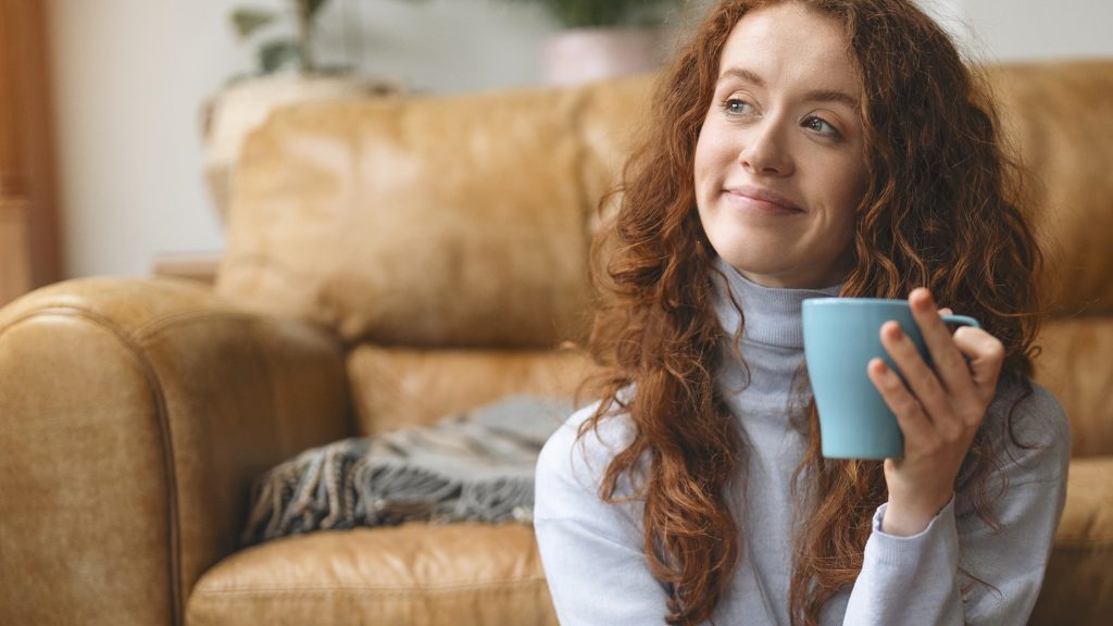 A young woman with curly hair holds a steaming mug, gazing into the distance, surrounded by a warm, inviting atmosphere. (Showcase file photo)